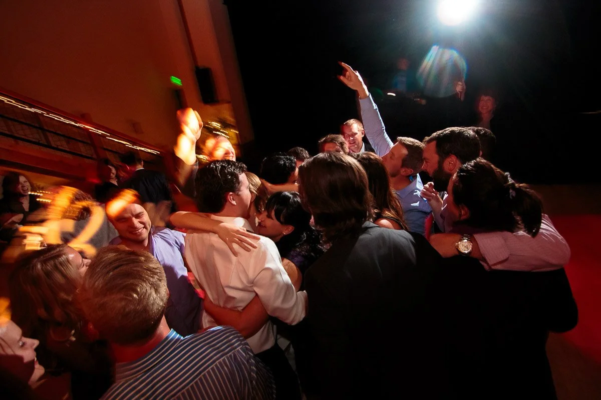 A lively group of people are gathered closely on a dance floor, embracing and celebrating. One person raises an arm triumphantly. Bright stage light shines above.