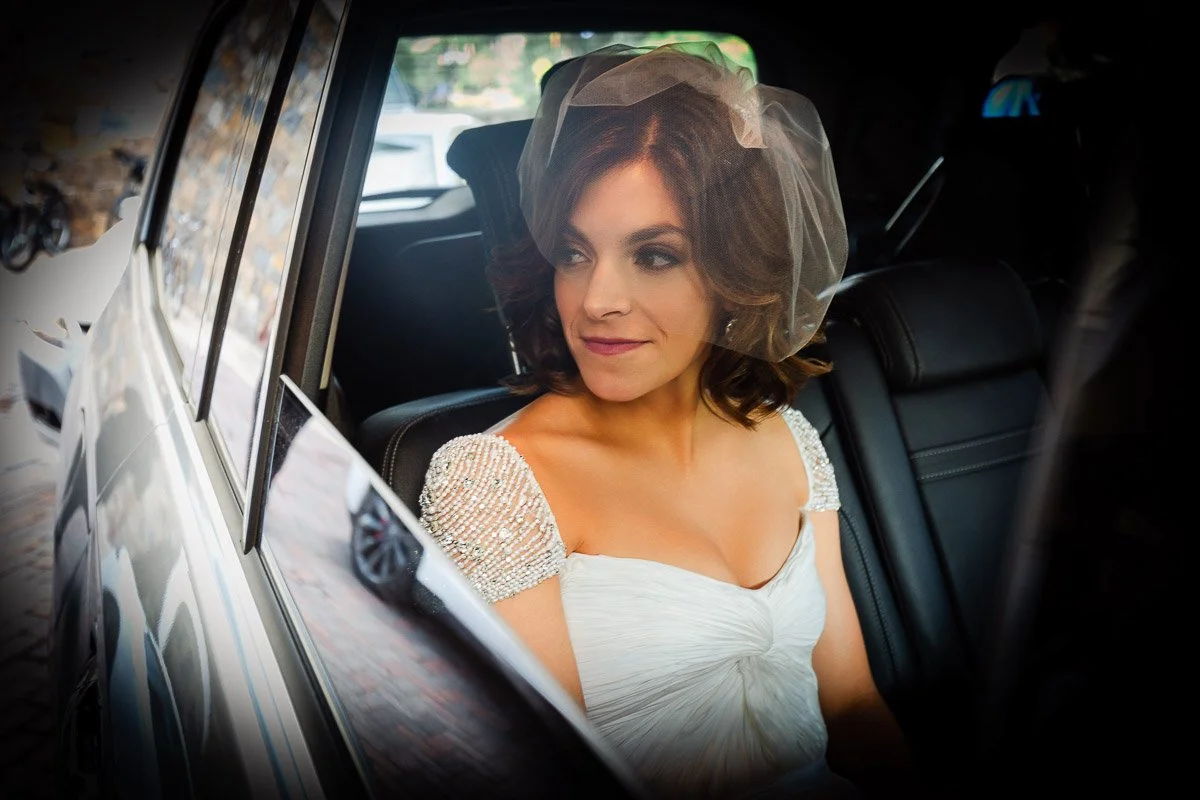 A bride in a beaded gown and veil looks out a car window with a serene expression. The interior is dimly lit, suggesting a calm, reflective moment.