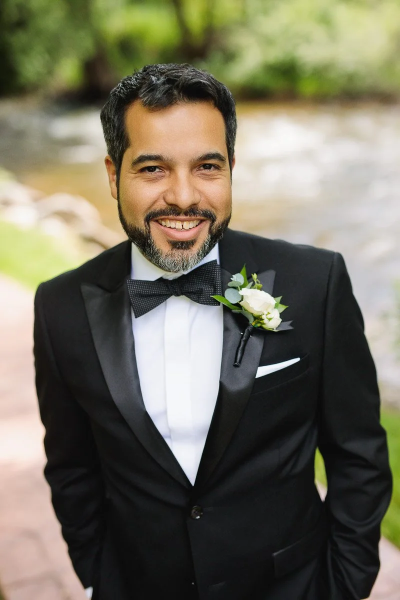 Smiling man in a black tuxedo with a white rose boutonniere stands on a garden path, exuding a joyful and relaxed vibe. Blurred greenery background.