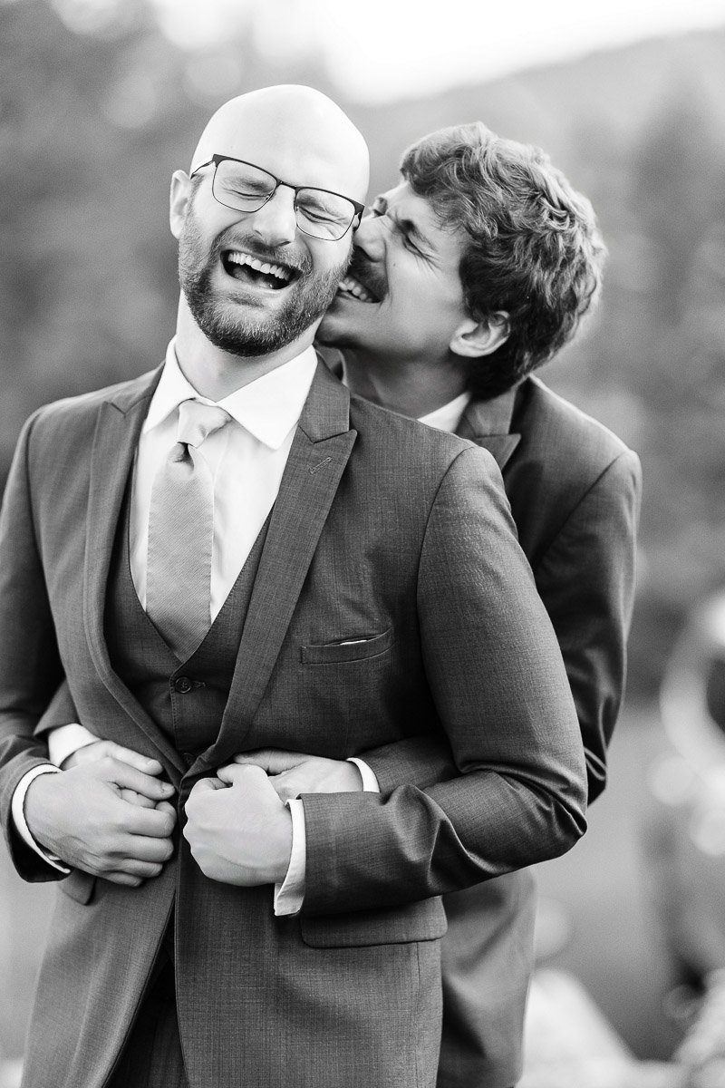 Two men in suits share a joyful moment, laughing and embracing. The affectionate scene conveys warmth and happiness against a blurred outdoor background.