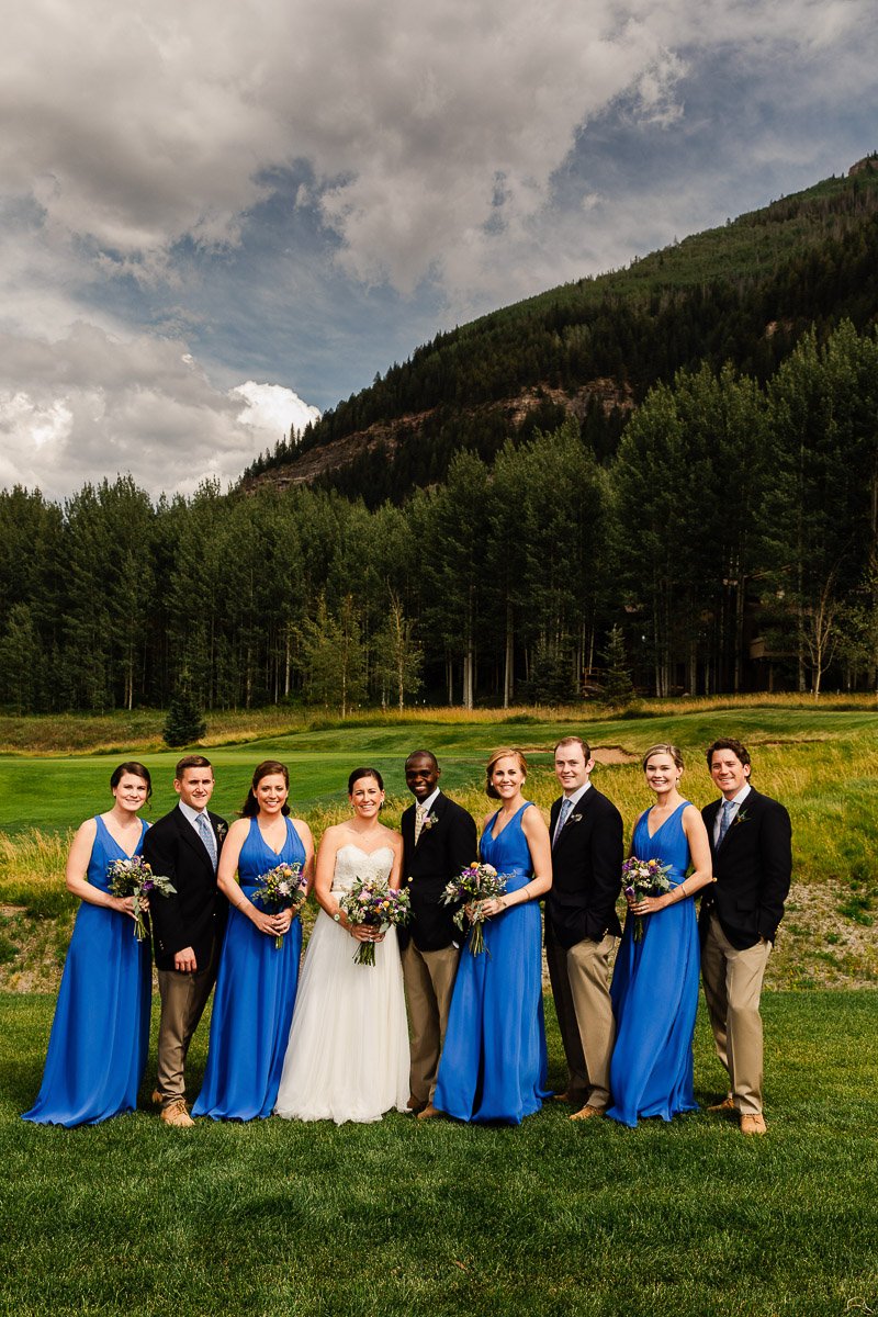 A joyful wedding party poses on a lush green lawn, backed by tall trees and a cloudy sky. The bride wears white, bridesmaids in blue.