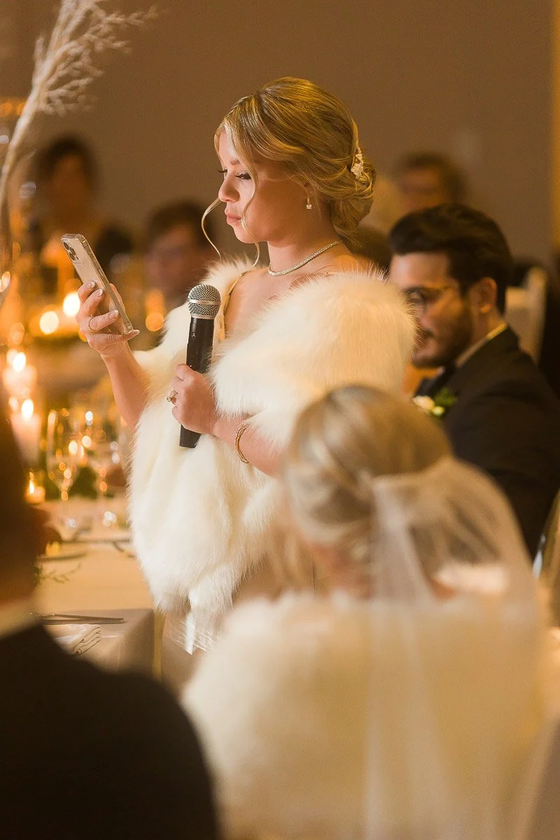 A woman in a white fur stole gives a speech at a wedding, holding a microphone and phone. Soft lighting and guests create a warm, intimate atmosphere.
