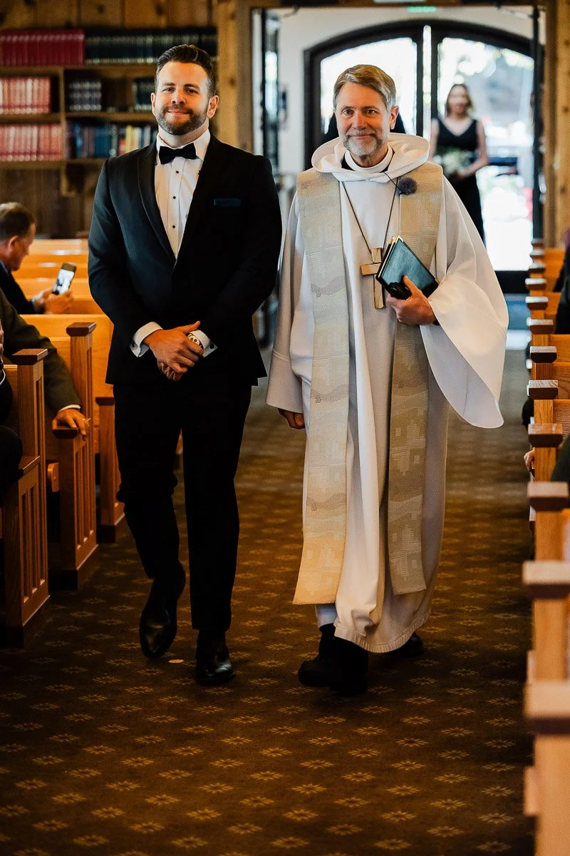 A smiling groom in a tuxedo walks with an officiant in robes down a church aisle. Guests watch them. The scene is warm and joyful.