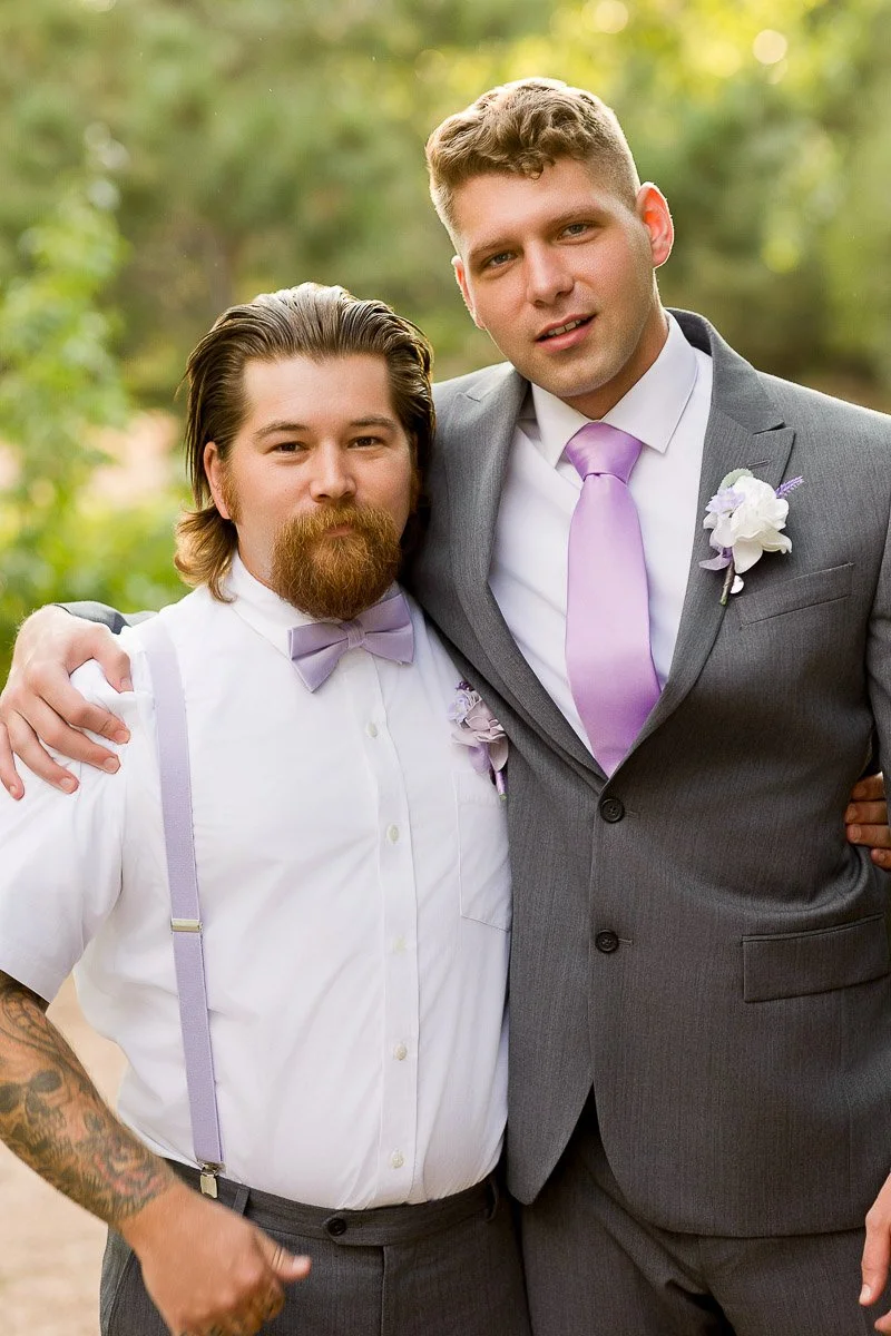 Two men smiling, standing arm-in-arm outdoors. The one on the left wears a white shirt with lavender suspenders; the other, a gray suit with a lavender tie.