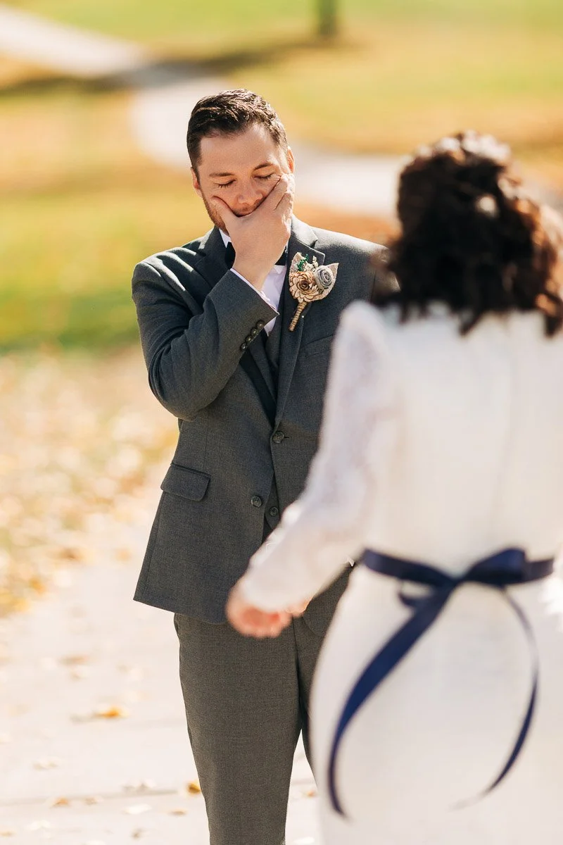 Groom in gray suit, visibly emotional with hand over mouth, sees bride in white dress with blue ribbon; autumn leaves scattered on the path.