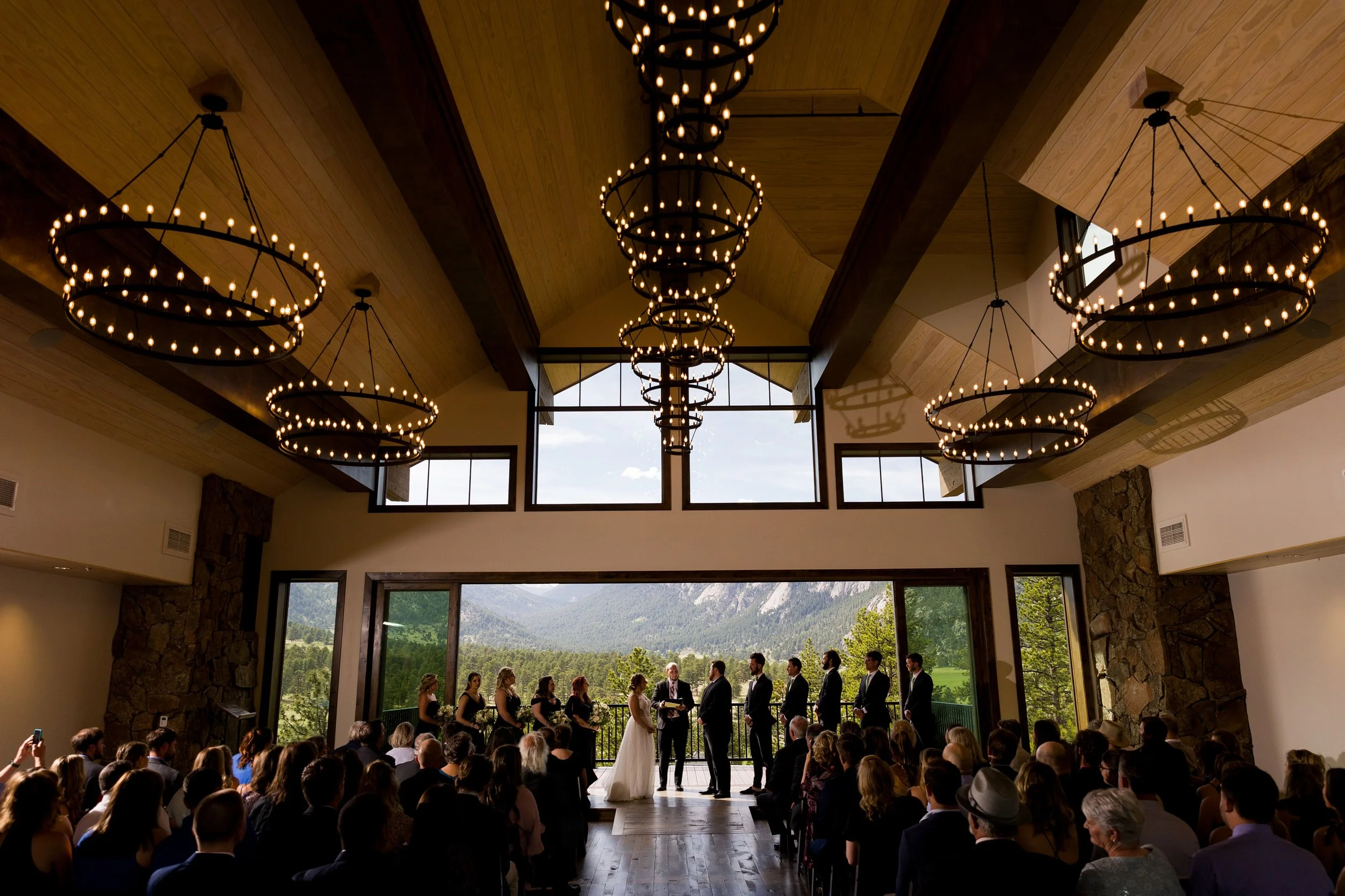 View a large wedding ceremony indoors with a full open wall section to the outside showing the Lumpy Ridge mountain range at Black Canyon Inn in Estes Park, Colorado