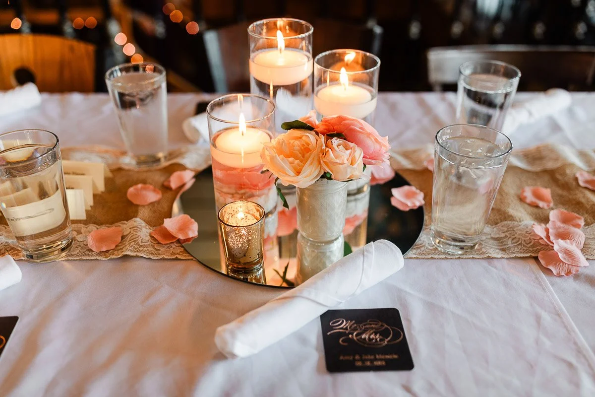 Elegant table setting with floating candles and peach roses on a mirrored centerpiece, surrounded by glasses, napkins, and pink petals, creating a romantic ambiance.