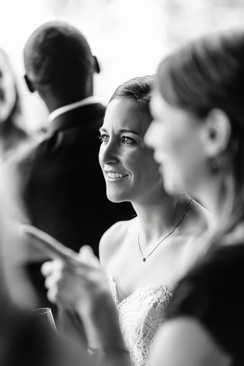 Smiling woman in focus, wearing a strapless white dress, surrounded by blurred people in formal attire at a social event; atmosphere is joyful and elegant.