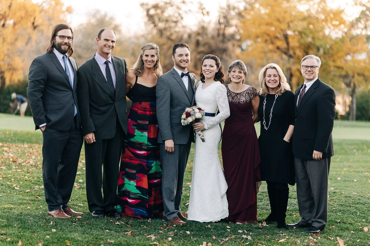 A group of eight people smile outdoors on a lawn with autumn trees in the background. A couple in wedding attire stands in the center, surrounded by loved ones.