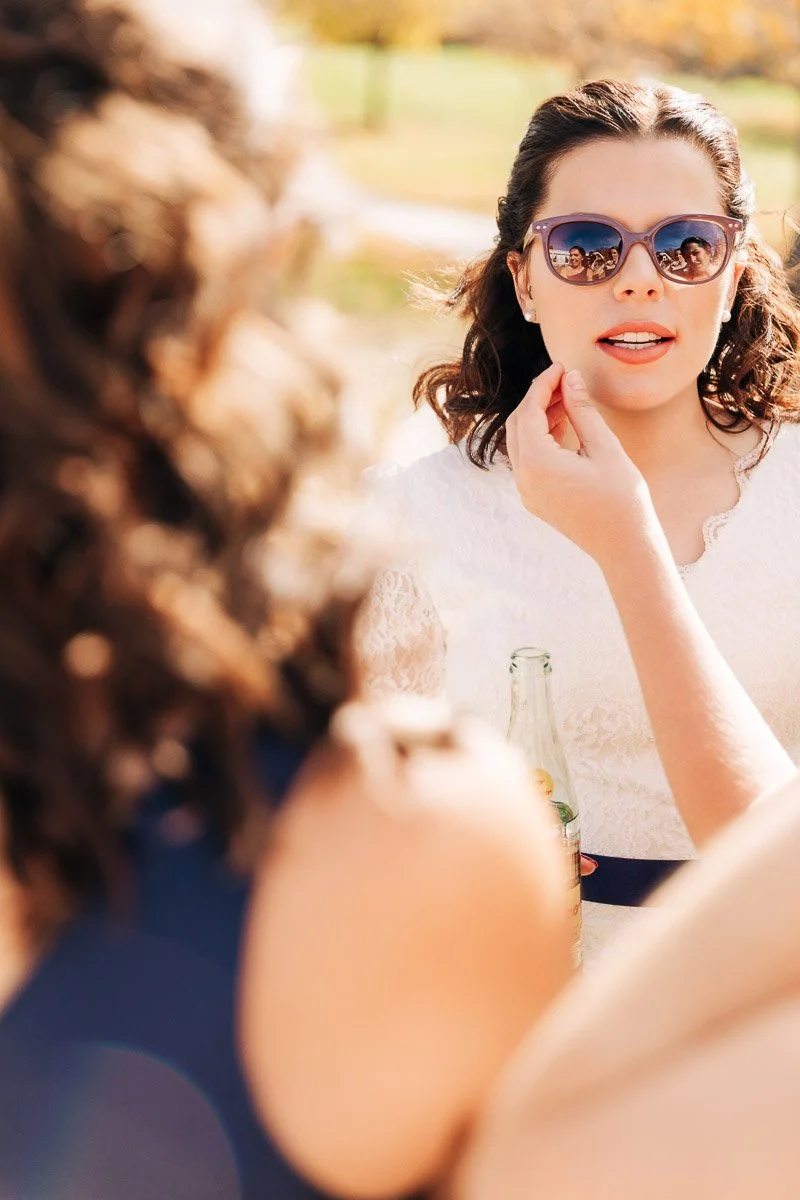 Bride in a lacy dress, wearing sunglasses, looks relaxed holding a drink. Her expression is calm as she talks to someone outside. Sunlit background.