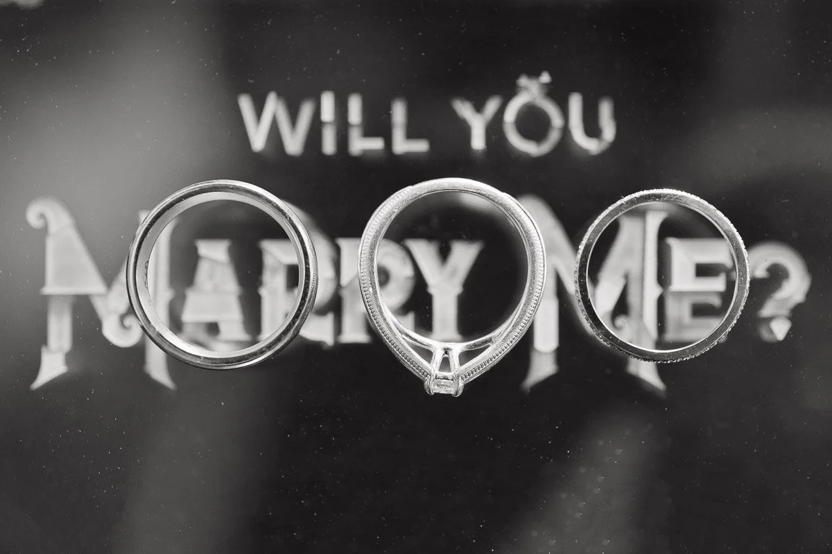 Three rings placed over the words "Will You Marry Me?" in a stylized font. The arrangement conveys a proposal theme with elegance and romance.