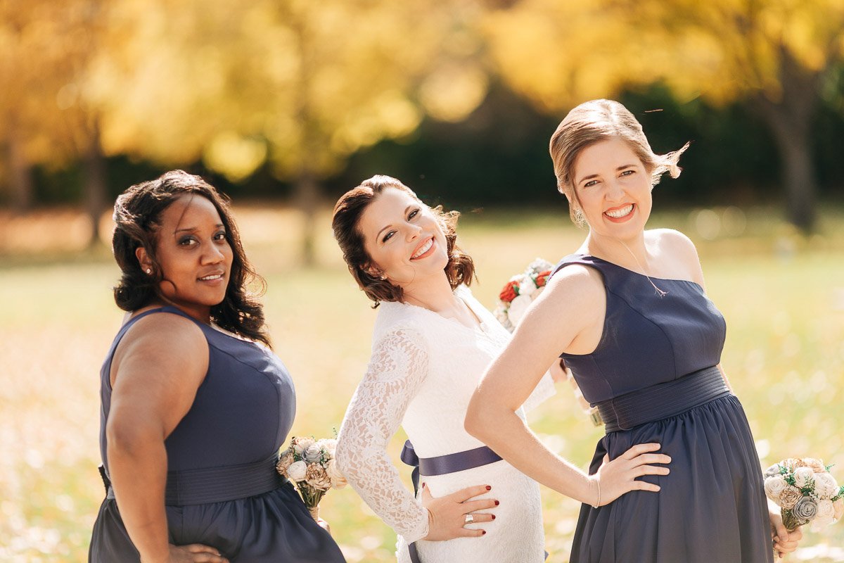 Three women smiling in a sunny park, wearing blue and white dresses. The background has autumn foliage, creating a warm, joyful atmosphere.
