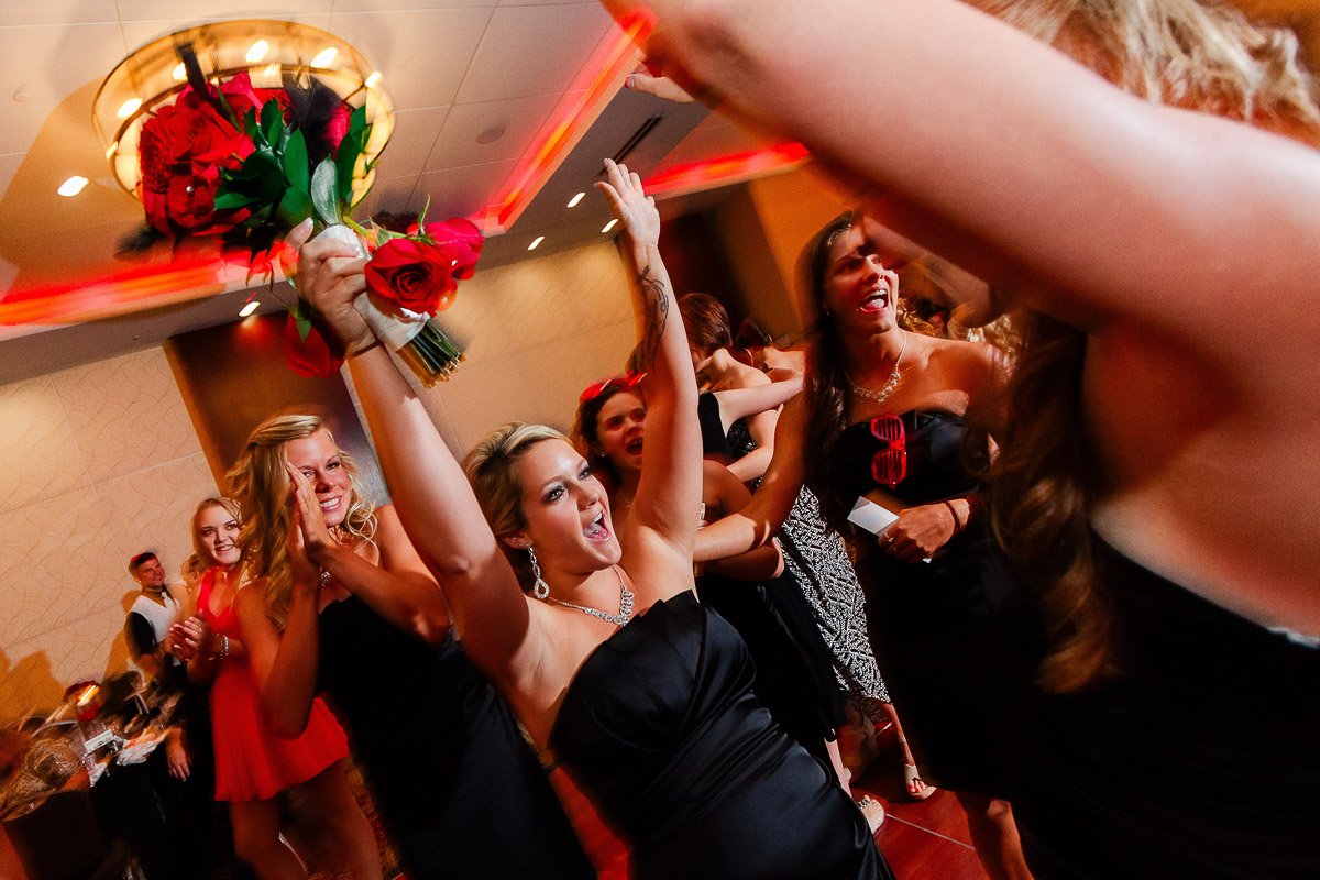 A joyous woman in a black dress holds a red rose bouquet overhead, surrounded by celebrating friends in an elegant, warmly lit venue.