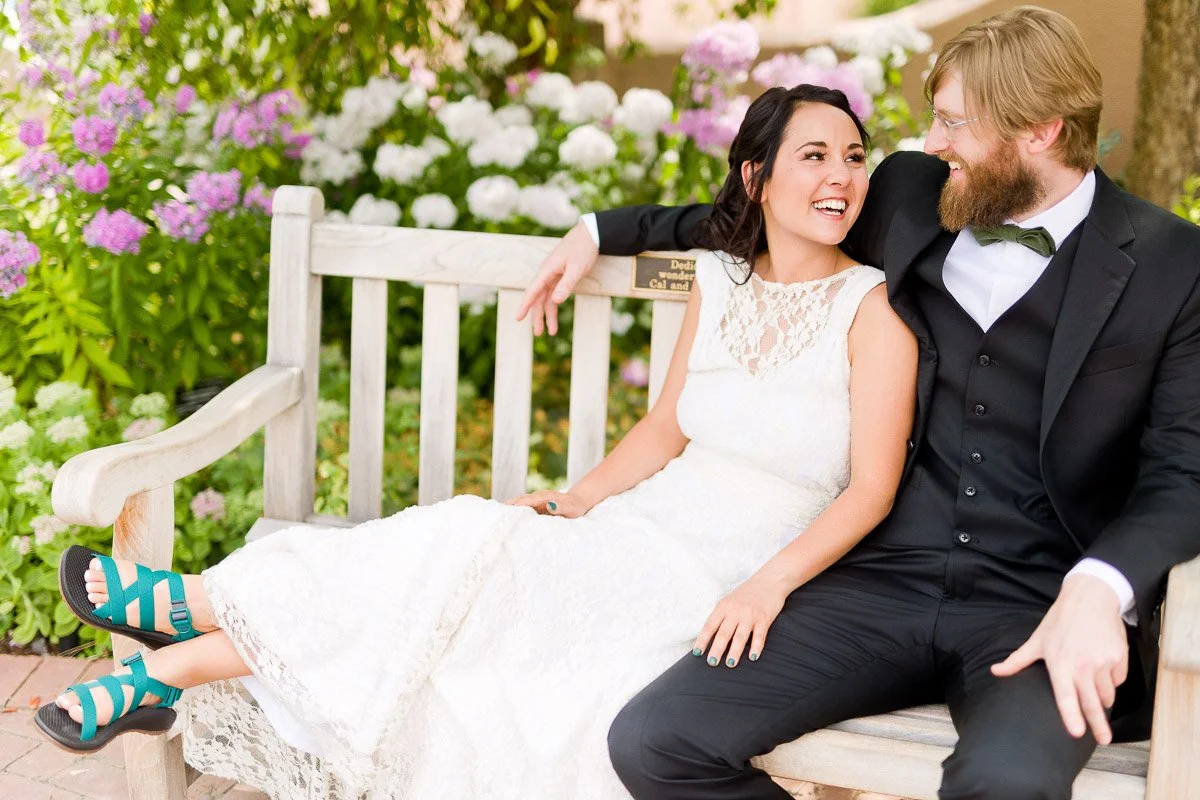 A couple in wedding attire sits on a wooden bench in a garden, laughing and looking at each other. The bride wears turquoise sandals, and flowers bloom behind them.