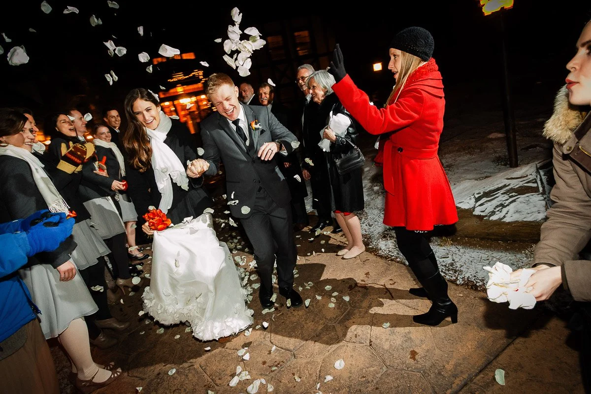 Bride and groom in a snowy outdoor setting, joyfully walking through a shower of flower petals, surrounded by smiling guests in winter attire.