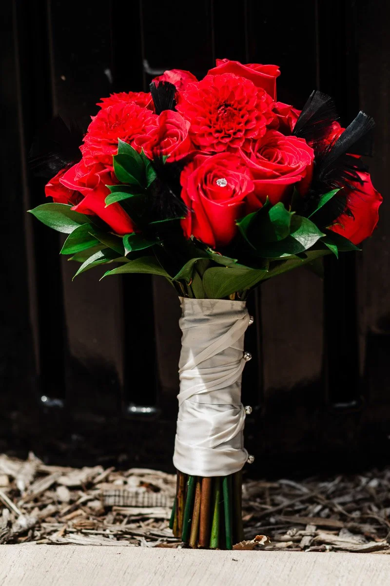 A bouquet of vibrant red roses and dahlias with black feathers, wrapped in white ribbon, rests against a dark backdrop, conveying a dramatic and elegant tone.