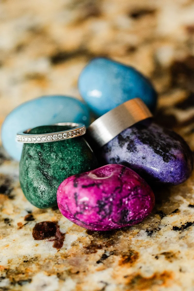 Colorful stones in blue, green, purple, and pink with two silver rings rest on a textured, speckled surface. The scene conveys a sense of elegance.