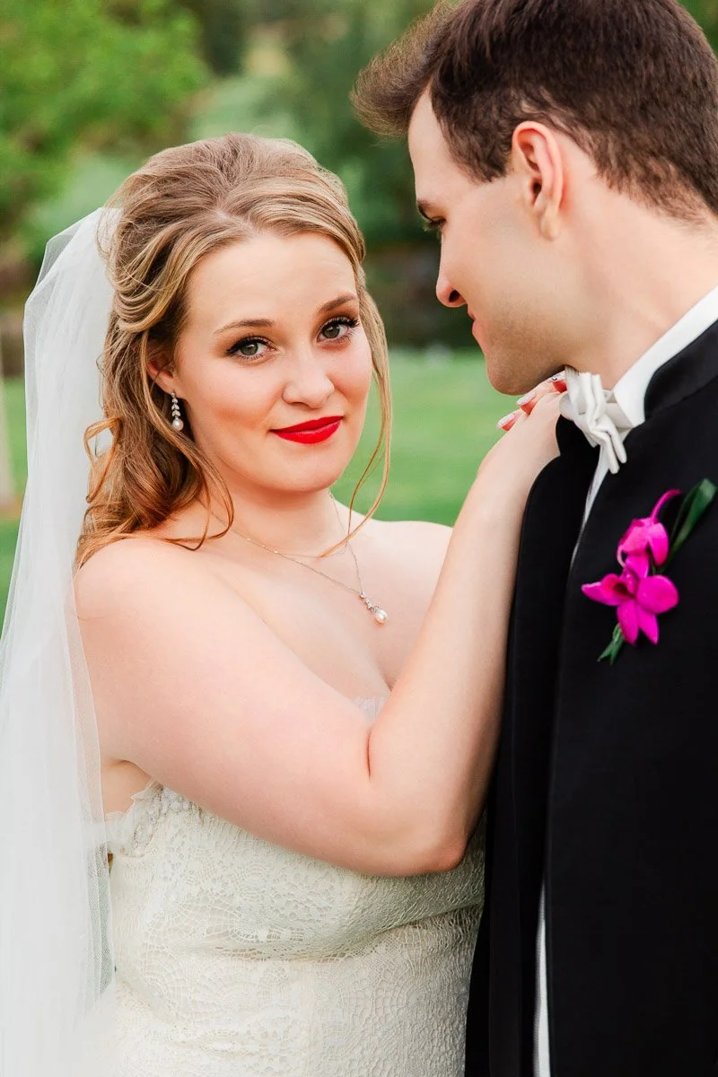Bride in white dress and veil smiles at the camera, touching the groom's shoulder. The groom wears a suit with a purple boutonnière, looking at her lovingly.