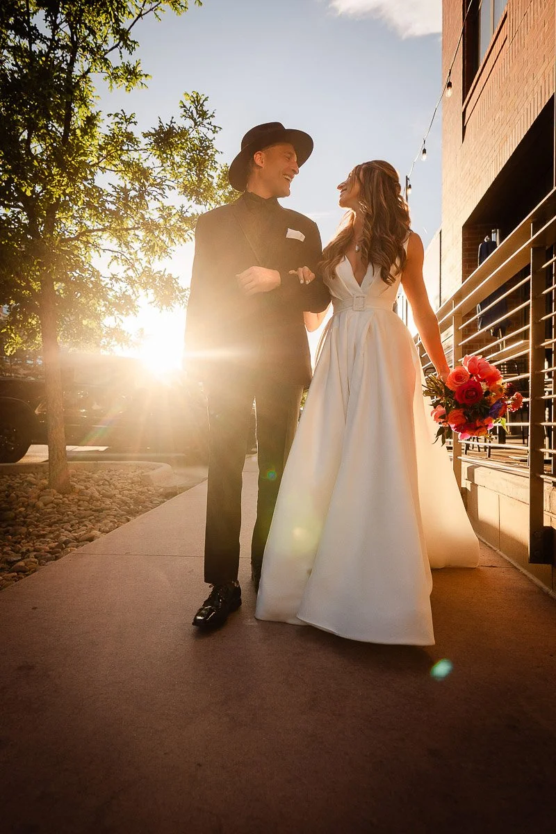 A couple walks arm in arm on a sunlit path, with the sun setting behind them. The woman wears a white gown, holding vibrant flowers, while the man is in a dark suit and hat.