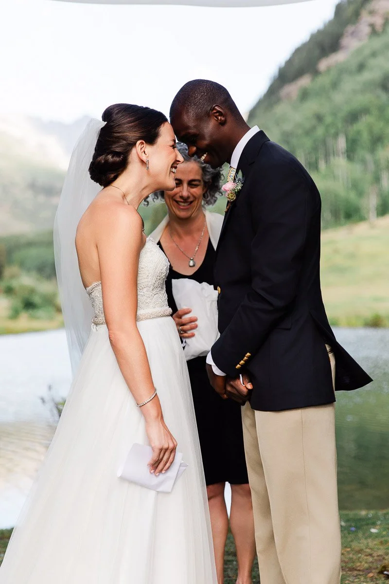 A bride in a white gown and groom in a dark suit share a joyful moment during their outdoor wedding ceremony, with a smiling officiant nearby.
