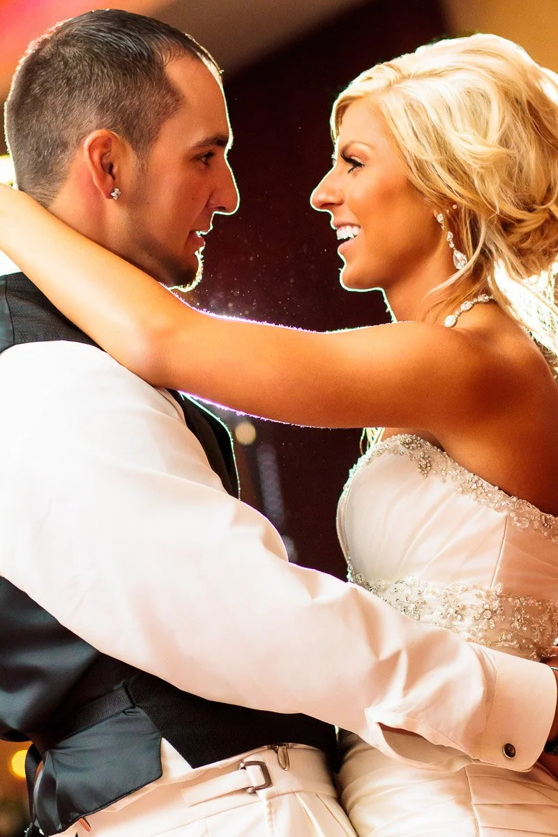 A bride and groom share a joyful dance, gazing into each other's eyes. The bride wears a strapless gown with sparkling details, while the groom dons a white shirt and vest.