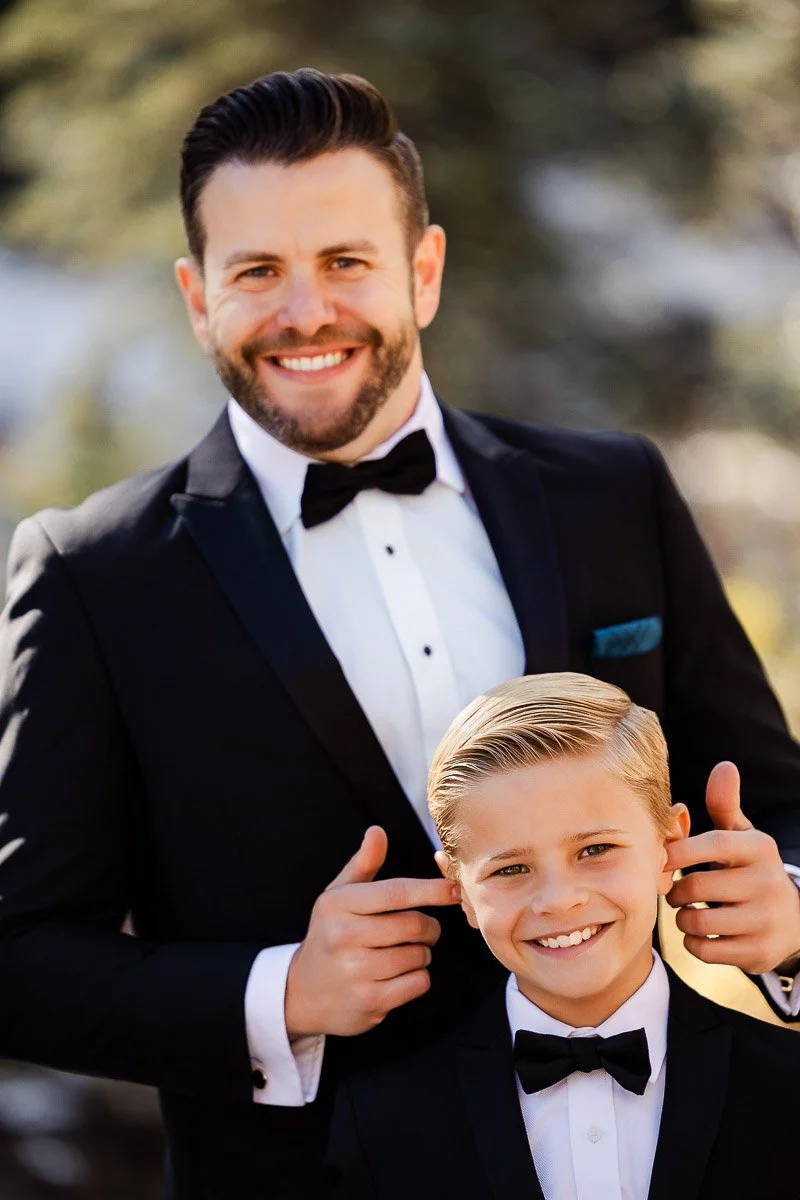 A smiling man and a boy, both in black tuxedos with bow ties, stand outdoors. The man gently holds the boy's shoulders, conveying a warm, joyful bond.