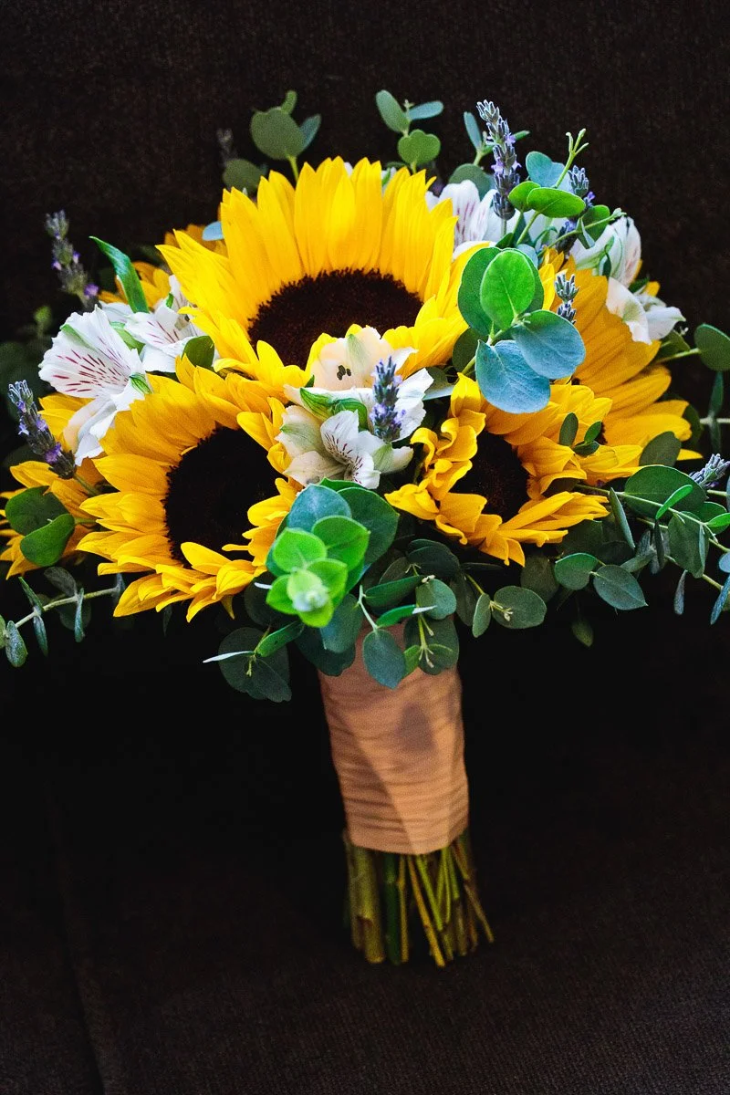 A vibrant bouquet features large sunflowers with dark centers, surrounded by white flowers, green foliage, and lavender sprigs, creating a cheerful, rustic feel.