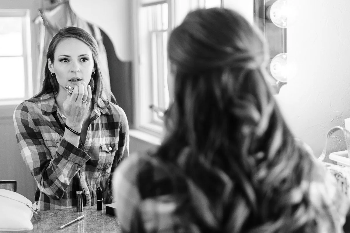A woman in a plaid shirt applies lipstick while looking in a mirror in a softly lit room. Her expression is focused and thoughtful.