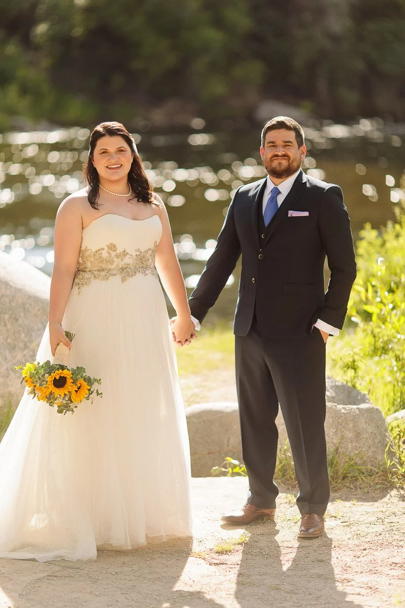 Bride and groom holding hands by a sunny riverside. She wears a white gown, holding sunflowers, and he wears a black suit with a blue tie. Romantic scene.