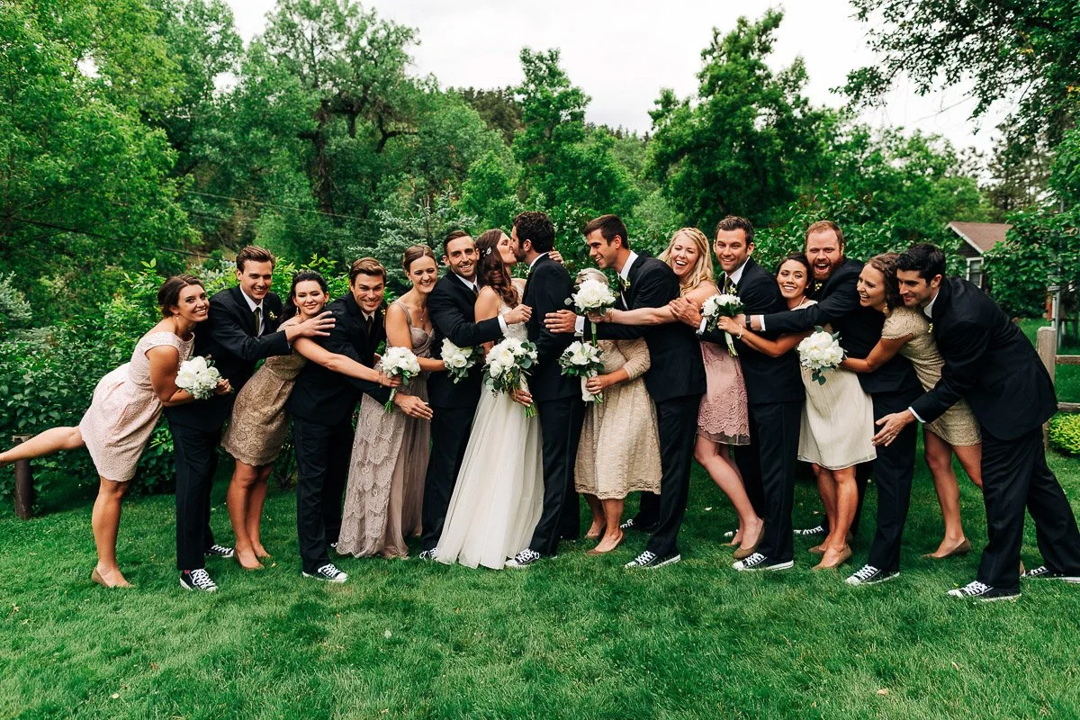 A joyful bridal party poses outdoors. The couple kisses in the center, surrounded by bridesmaids in beige dresses and groomsmen in suits, all wearing matching sneakers.