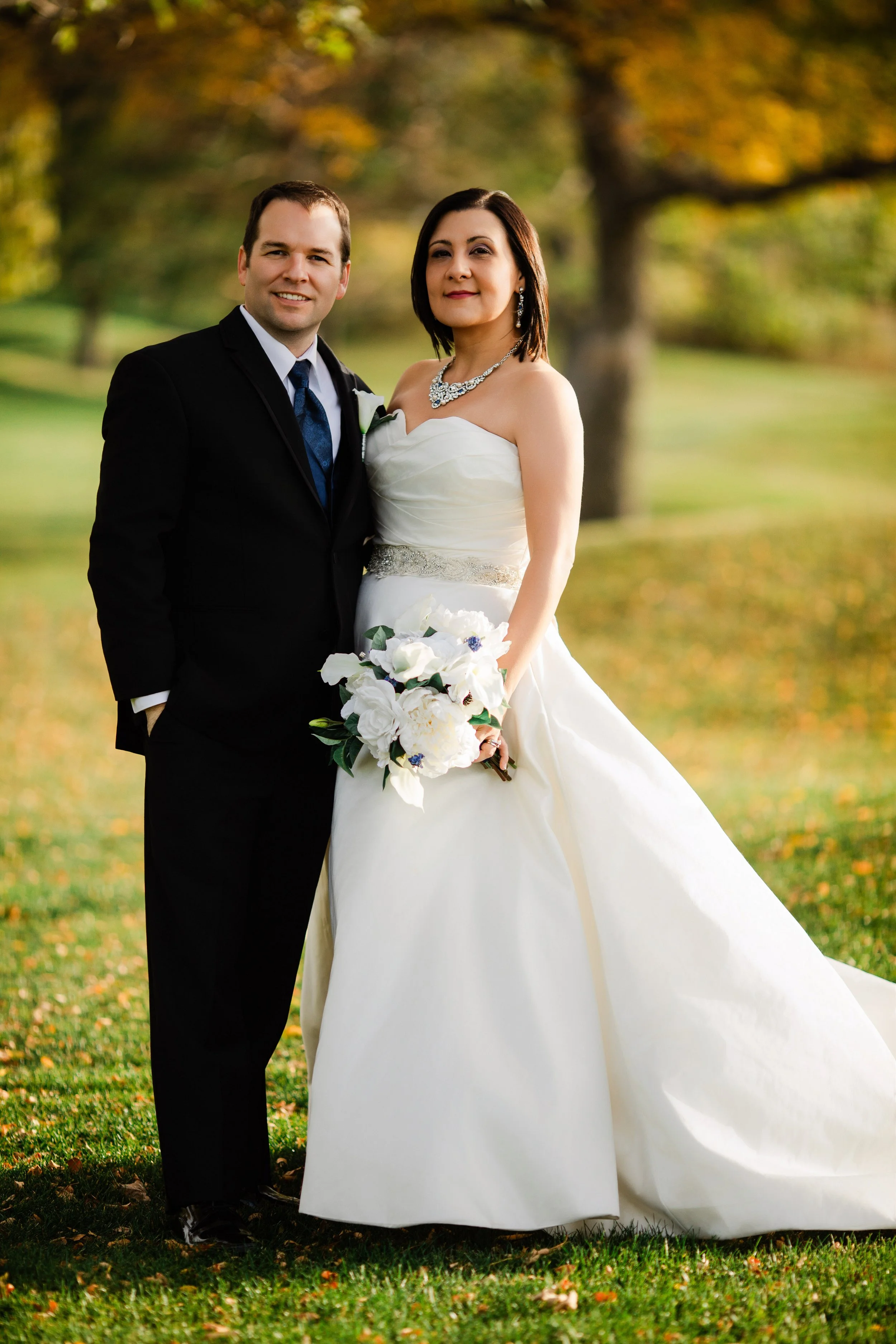 Bride and groom smiling outdoors. The bride wears a white gown with a bouquet; the groom is in a dark suit. Sunlit autumn leaves in the background.