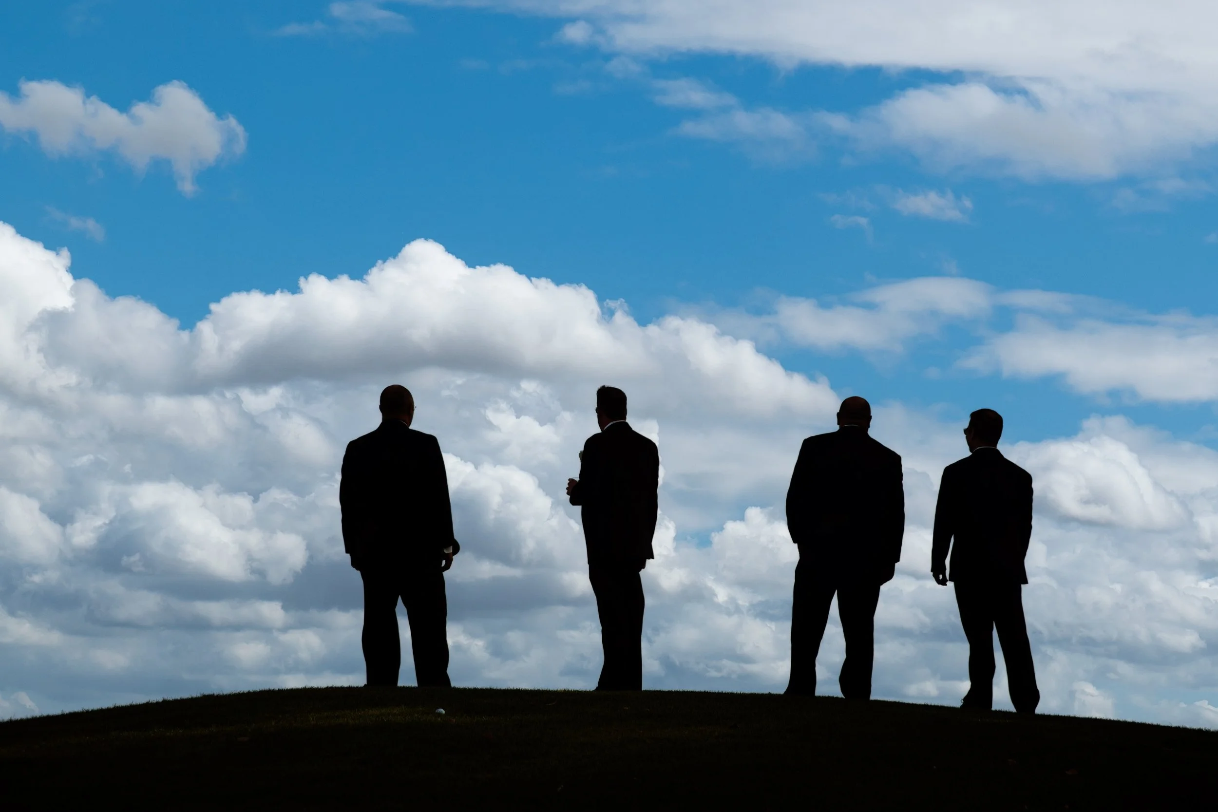 Silhouetted against a bright blue sky with fluffy clouds, four men in suits stand on a hill, conveying an air of professionalism and contemplation.