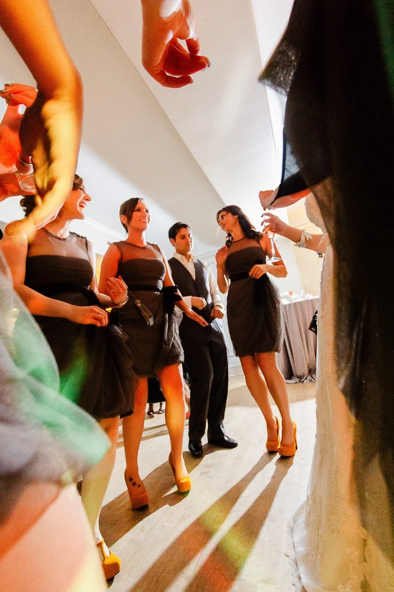 A lively wedding dance floor scene with people in elegant attire. Three women in matching dresses and a man in a suit are joyfully dancing together. Warm lighting creates a festive atmosphere.