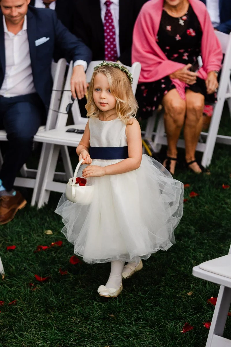 A young girl in a white dress and flower crown walks down an aisle, holding a basket of red petals. Guests in formal attire watch her, seated on white chairs.
