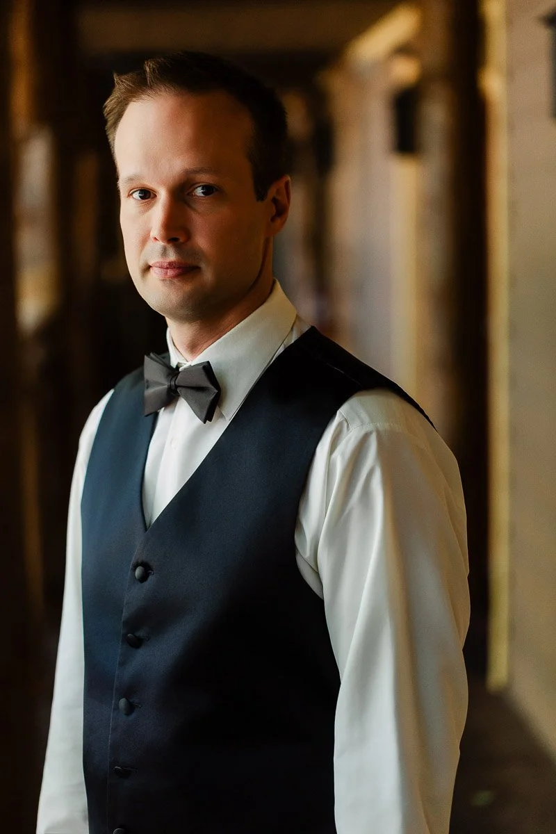 A man in formal attire stands indoors, wearing a black vest, white shirt, and black bow tie. He gazes calmly at the camera, background softly blurred.