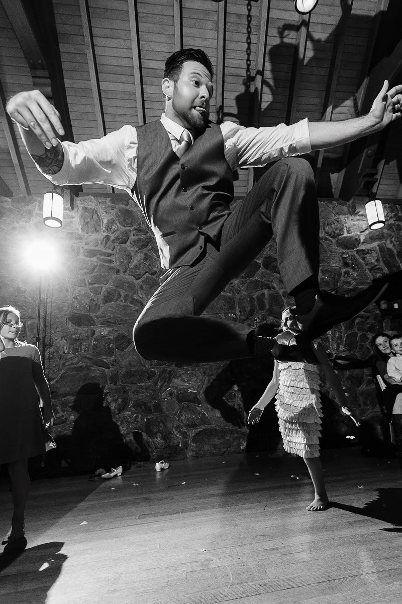 A man in a suit jumps energetically in a dimly lit room with a stone wall. Guests watch in the background, creating a festive, joyful atmosphere.