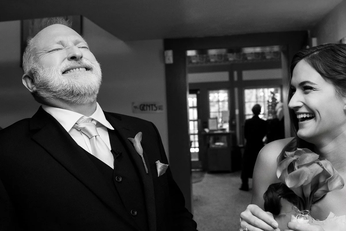 A bearded man in a suit and a woman in a dress share a joyful laugh in a hallway. The woman holds a bouquet, exuding a warm and lighthearted mood.