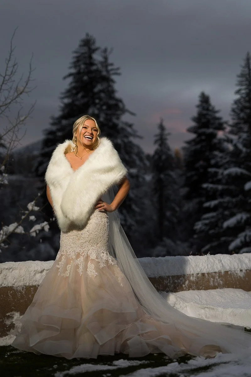 A joyful woman in a wedding gown and white fur shawl stands in a snowy landscape with pine trees. The sky is overcast, conveying a serene, wintry atmosphere.