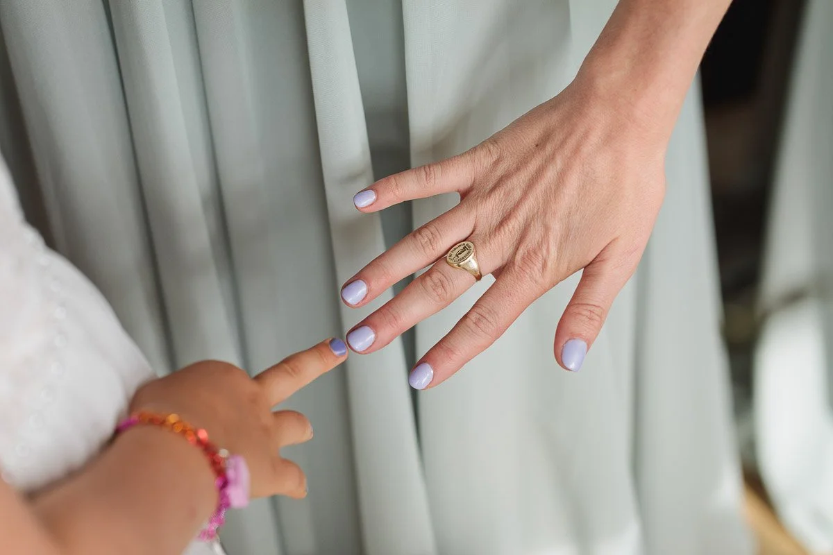 A child and adult with light purple nails touching fingertips, conveying a sense of connection. The adult wears a gold ring. Both wear light clothing.