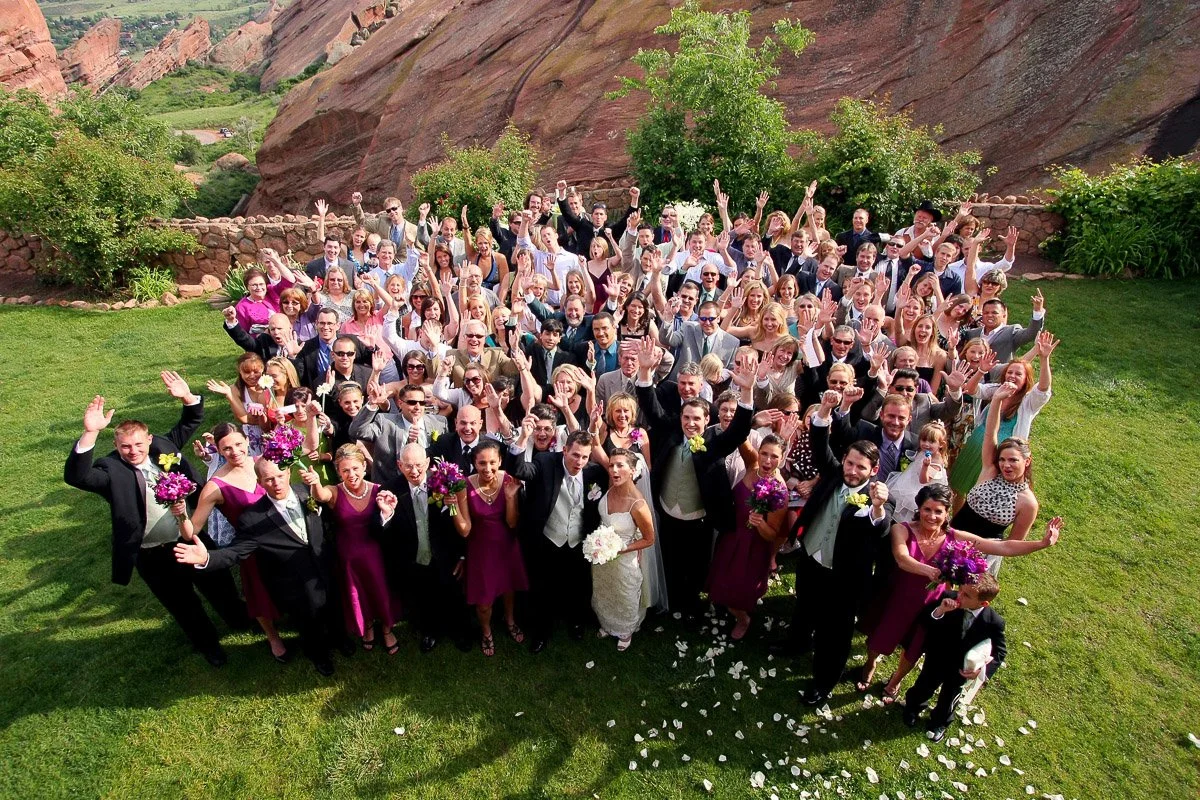 A joyful wedding group photo with many guests, mostly dressed formally, waving and smiling on a green lawn with red rock formations in the background.