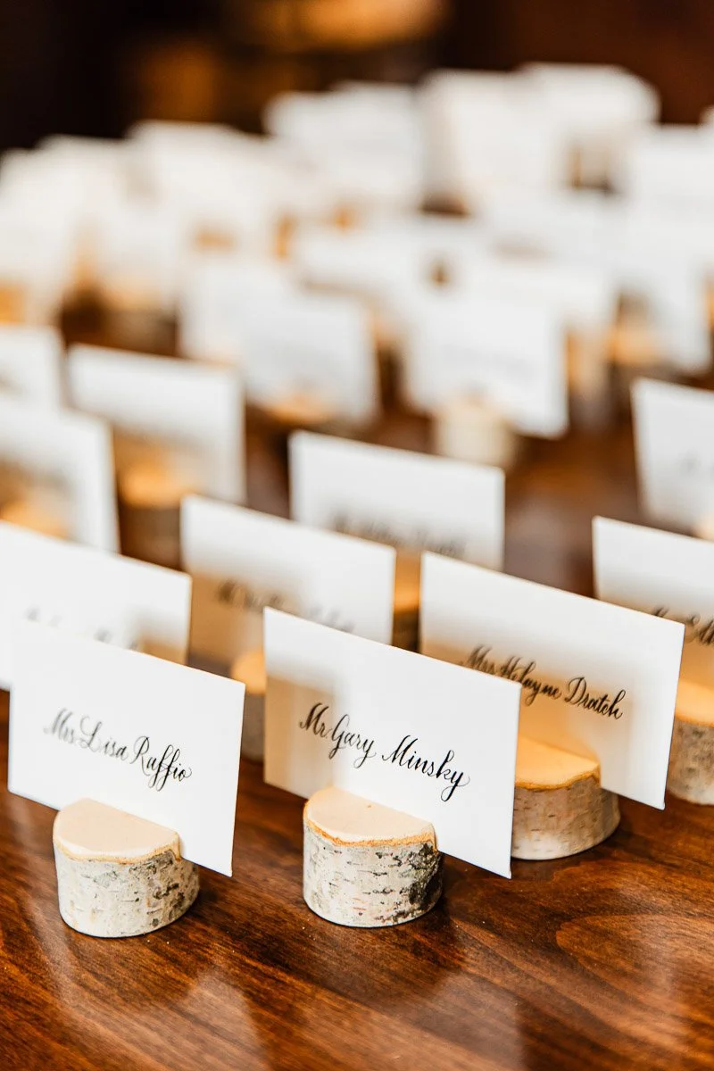 Rows of elegant white place cards with cursive writing are held by birch wood holders on a polished wooden table, creating a warm and inviting tone.