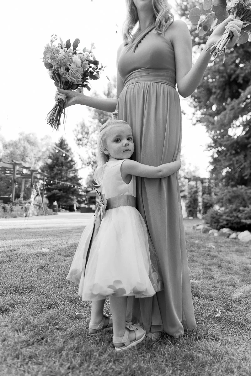 A young girl in a dress hugs a woman's leg, who holds two bouquets. They stand on grass, with trees in the background, creating a serene scene.