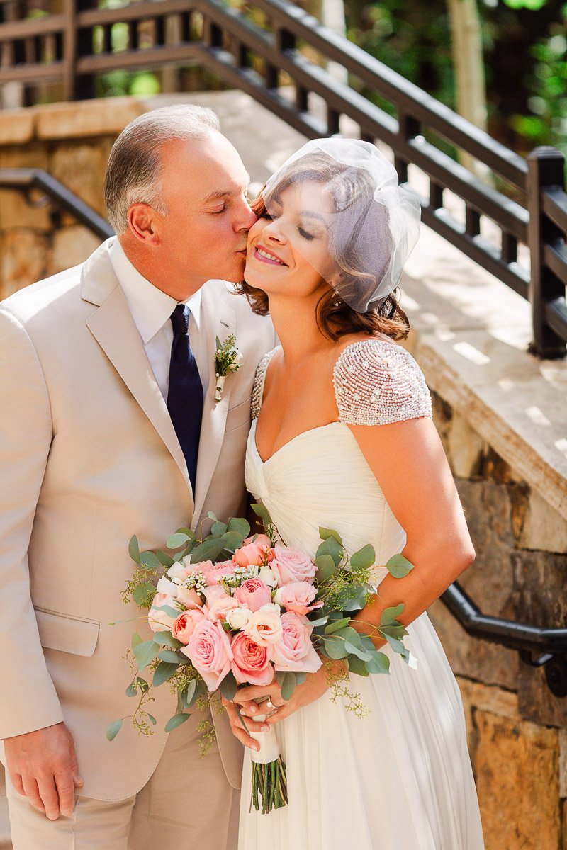 Bride and groom share a joyful embrace. She holds pink roses, and he kisses her cheek. Both are elegantly dressed, creating a warm, romantic atmosphere.