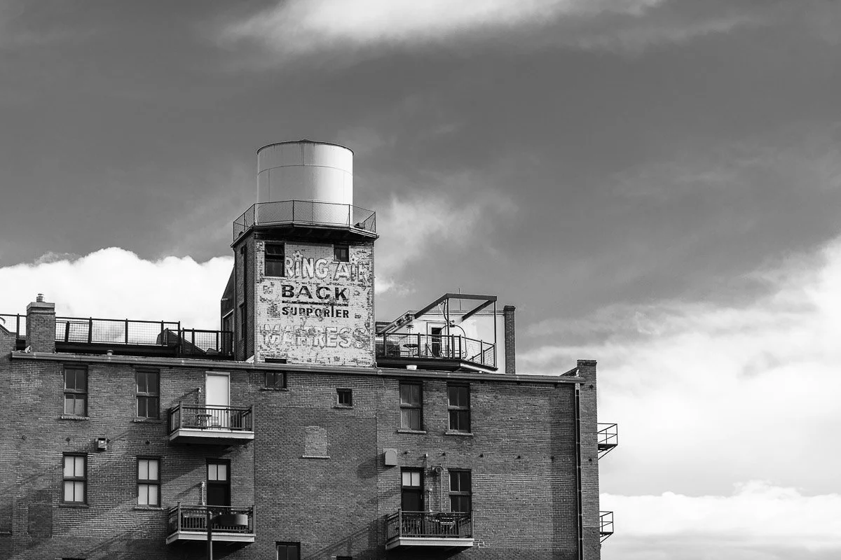 Black and white image of a brick building with a water tower. Faded advertisement on the wall, dramatic clouded sky above, evokes nostalgia.