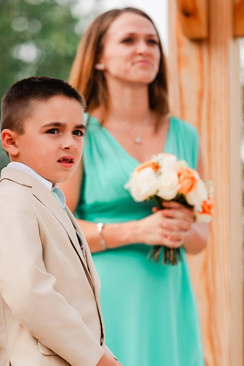 A young boy in a beige suit looks to the side with a curious expression. Behind him, a woman in a teal dress holds a bouquet of orange and white flowers. The setting is outdoors, conveying a formal, joyful atmosphere.