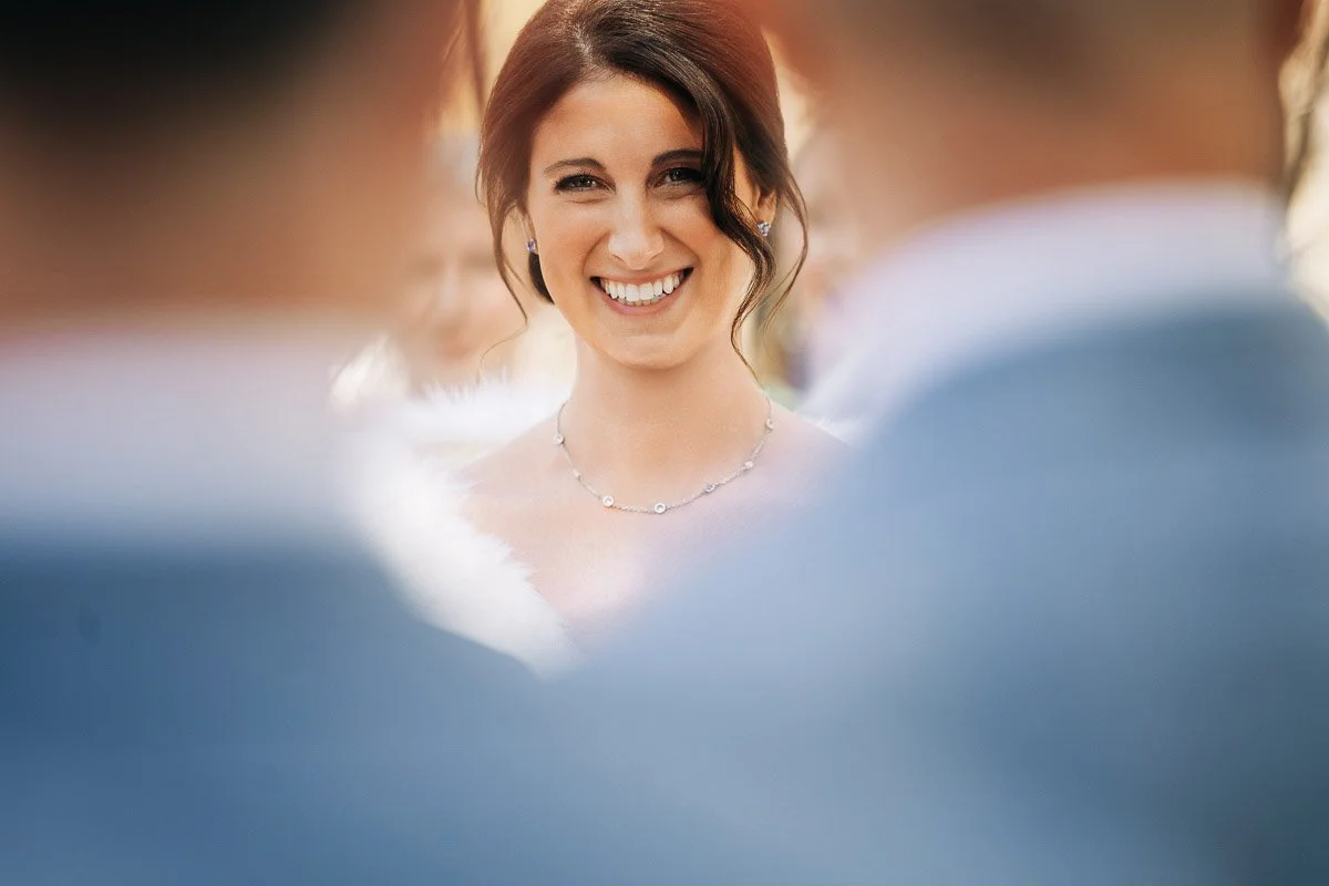 A smiling bride in white with an elegant necklace gazes warmly, framed by two blurred figures in blue suits, conveying joy and celebration.