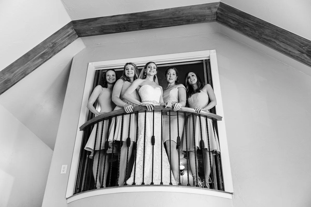 A bride in a strapless gown stands with four bridesmaids on a wooden balcony. All are smiling, conveying joy and togetherness in a wedding setting.