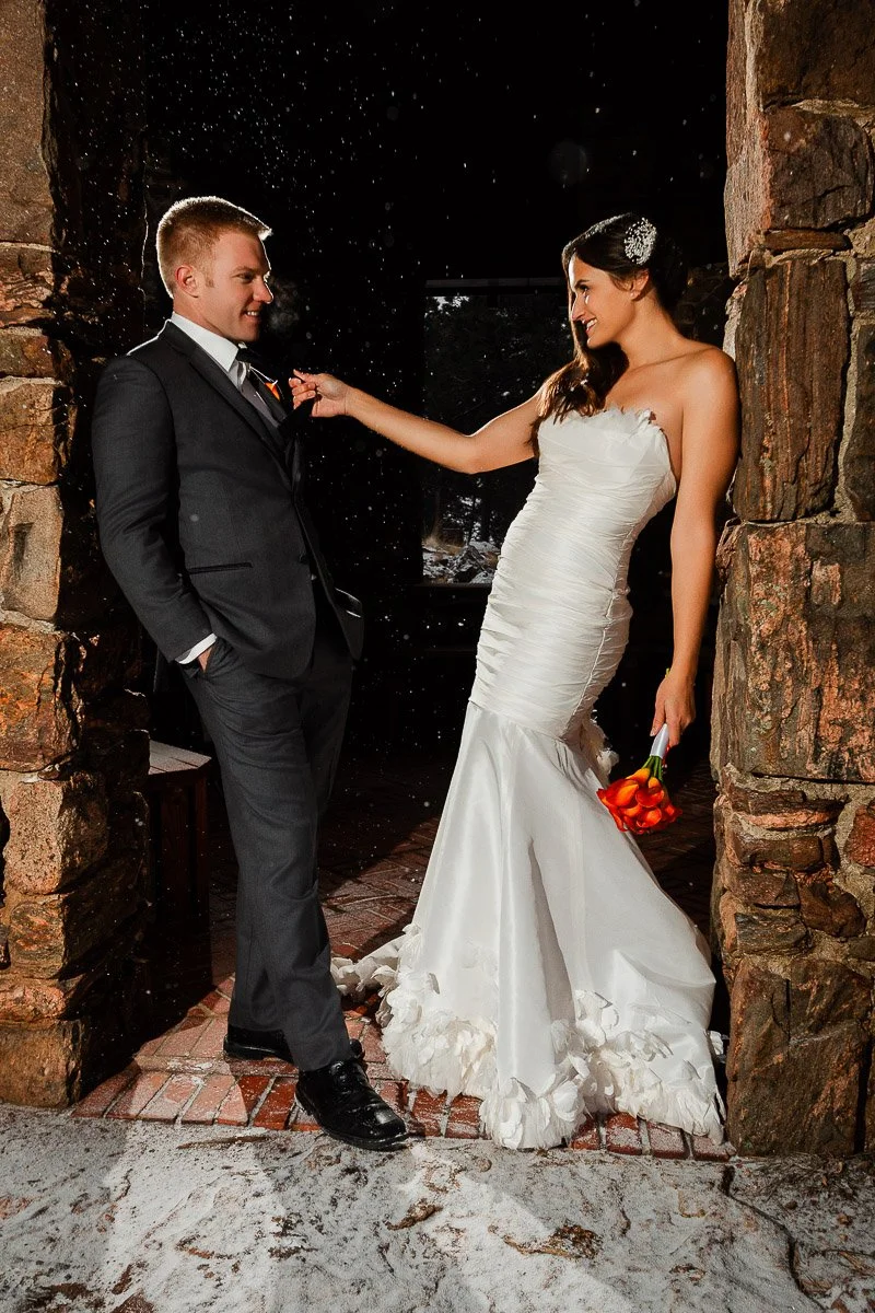 A bride in a white dress playfully adjusts a groom's tie in a dark stone archway during a Boettcher Mansion wedding. She holds orange flowers, and light snow covers the ground.
