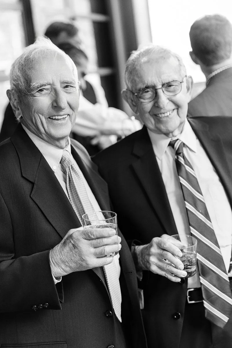 Two elderly men in suits smile while holding drinks at a social gathering. They stand close together, conveying a sense of companionship and joy. Black and white image.