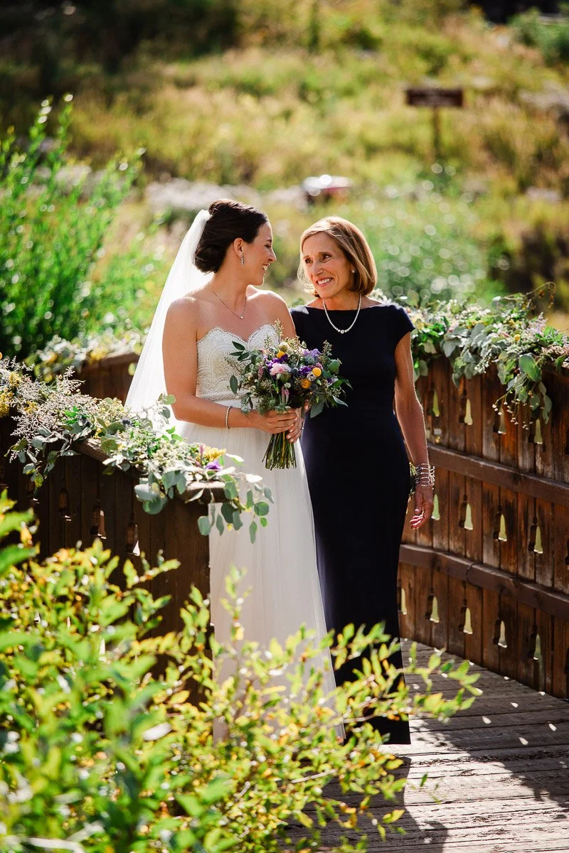 A bride in a white dress and veil walks with her Mother in a dark dress on a wooden bridge adorned with greenery, sharing a joyful moment during a Vail Golf Club wedding in Vail, Colorado